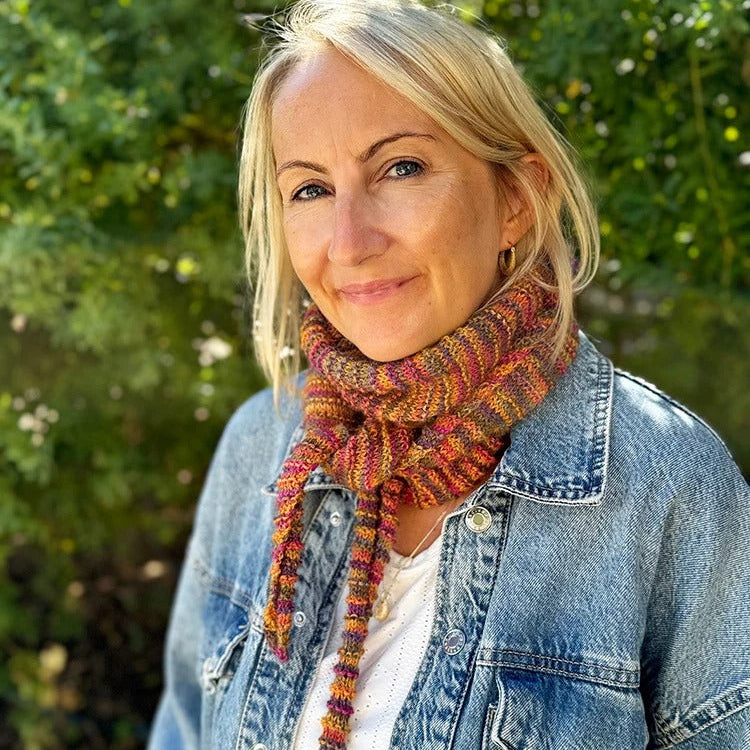 Woman wearing a colorful scarf and denim jacket outdoors with greenery in the background